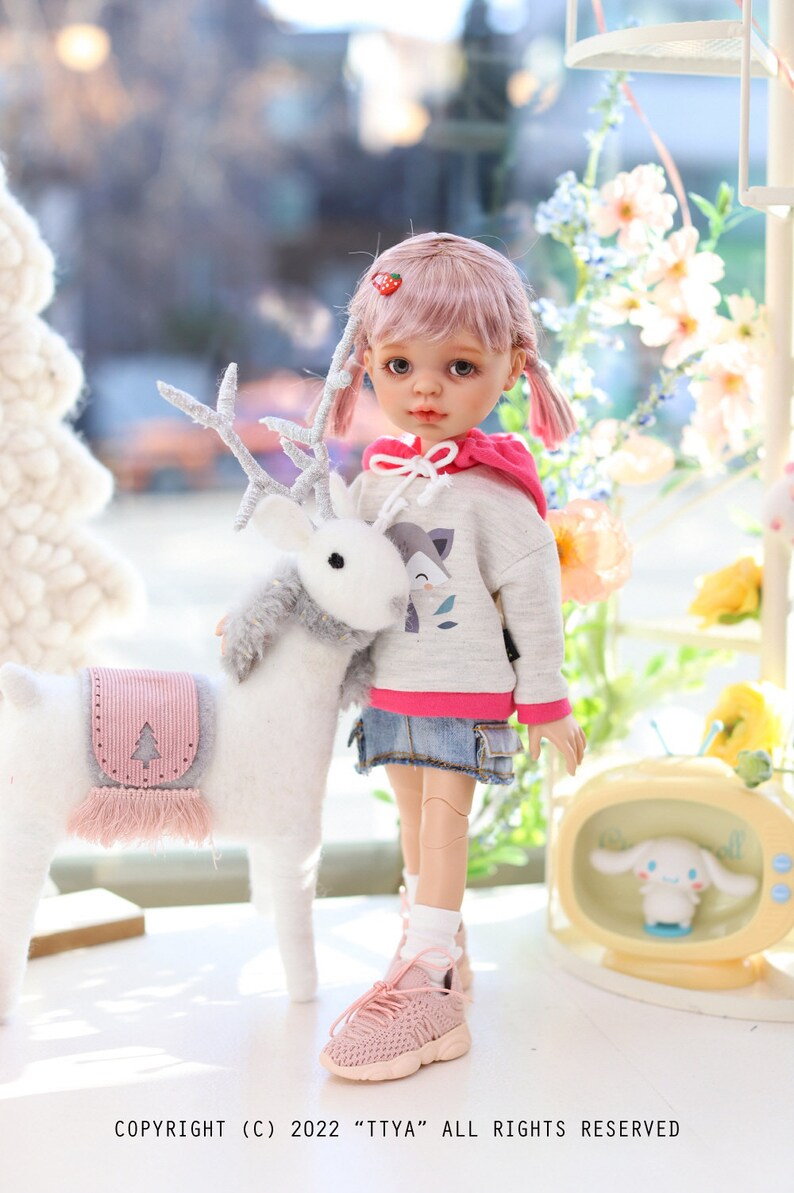 Pode incluir: Uma boneca rosa e cinza com cabelo rosa e tran&ccedil;as usando um moletom cinza com um gr&aacute;fico de raposa e uma saia jeans. A boneca est&aacute; segurando uma rena de pel&uacute;cia branca com chifres. A boneca est&aacute; usando t&ecirc;nis rosa.