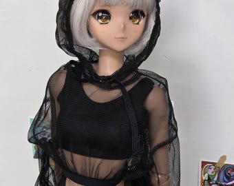 Puede incluir: Una muñeca con cabello plateado y ojos grandes que lleva una parte superior con capucha de malla negra, un top corto a juego y una falda. La muñeca posa sobre un fondo blanco, con impresiones artísticas visibles en la pared.