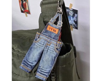 Puede incluir: Un llavero de estilo denim con forma de peto en miniatura, con el texto "TTYA" en la parte delantera. El llavero está sujeto a un cierre plateado y cuelga de un bolso verde. Dos pequeñas fotos están pegadas a la pared detrás del bolso.
