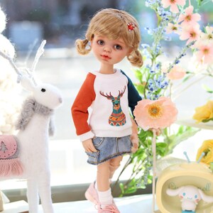 Pode incluir: Uma boneca com cabelo loiro em tran&ccedil;as usando um su&eacute;ter branco, laranja e turquesa com um design de veado. A boneca est&aacute; usando uma saia jeans e t&ecirc;nis rosa. A boneca est&aacute; em p&eacute; em frente a um fundo branco com flores.