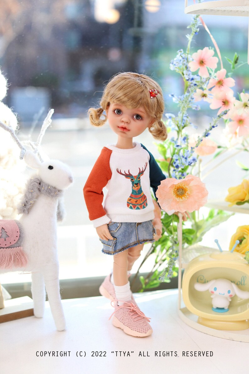 Pode incluir: Uma boneca com cabelo loiro em tran&ccedil;as usando um su&eacute;ter branco, laranja e turquesa com um design de veado. A boneca est&aacute; usando uma saia jeans e t&ecirc;nis rosa. A boneca est&aacute; em p&eacute; em frente a um fundo branco com flores.