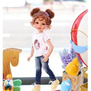 Puede incluir: Una mu&ntilde;eca con el pelo casta&ntilde;o en coletas que lleva una camiseta blanca con una imagen de una ni&ntilde;a, pantalones vaqueros azules y zapatillas amarillas. La mu&ntilde;eca est&aacute; de pie sobre una superficie de madera delante de una ventana. La mu&ntilde;eca est&aacute; mirando hacia la derecha de la imagen. COPYRIGHT (C) 2021 "TTYA" ALL RIGHTS RESERVED.