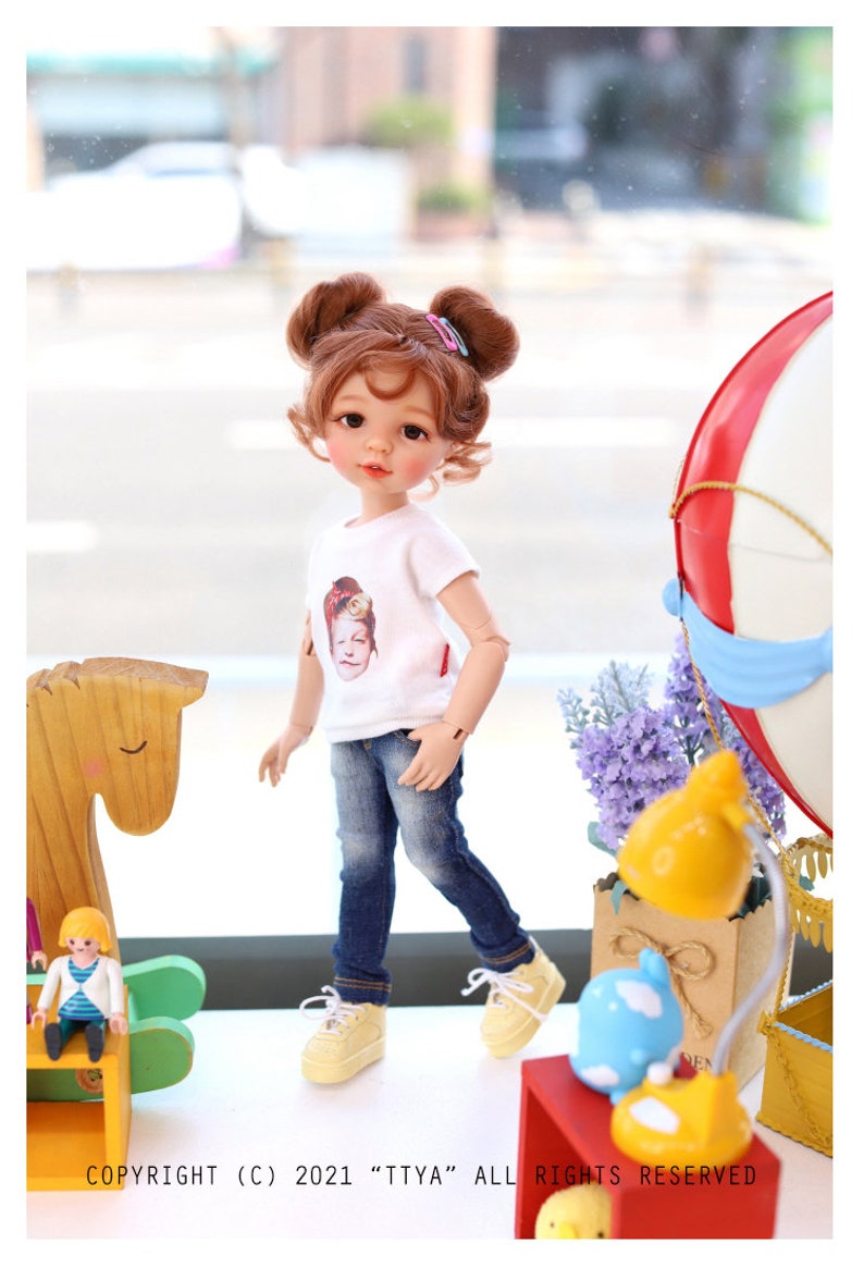 Puede incluir: Una mu&ntilde;eca con el pelo casta&ntilde;o en coletas que lleva una camiseta blanca con una imagen de una ni&ntilde;a, pantalones vaqueros azules y zapatillas amarillas. La mu&ntilde;eca est&aacute; de pie sobre una superficie de madera delante de una ventana. La mu&ntilde;eca est&aacute; mirando hacia la derecha de la imagen. COPYRIGHT (C) 2021 "TTYA" ALL RIGHTS RESERVED.