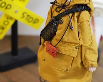 Puede incluir: Una muñeca con una chaqueta con capucha de color amarillo mostaza, una riñonera negra y una pequeña bolsa roja. La chaqueta tiene bolsillos grandes y detalles de botones metálicos. Un letrero amarillo con la palabra "STOP" está en el fondo.