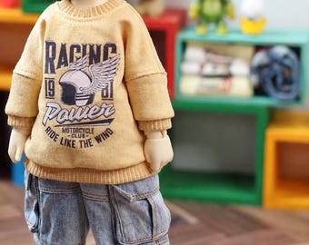 May include: A doll wearing a mustard yellow long-sleeve sweatshirt with a graphic print that reads Racing 1981 Power Motorcycle Club Ride Like The Wind and a winged helmet. The doll also wears light blue denim cargo shorts with visible pockets.