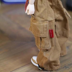 May include: Close-up of a doll's lower body, showcasing brown cargo pants with a large pocket on the leg. The pants have a red tag with white text. The doll is wearing white and tan sneakers. The doll's hand is visible in the pocket.