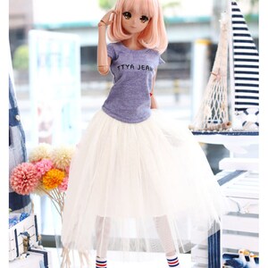 K&ouml;nnte beinhalten: Eine Puppe tr&auml;gt ein lila T-Shirt mit dem Aufdruck "TTYA JEAN", einen wei&szlig;en T&uuml;llrock, blau-rot gestreifte Socken und blaue Turnschuhe. Die Puppe hat rosa Haare und blaue Augen.