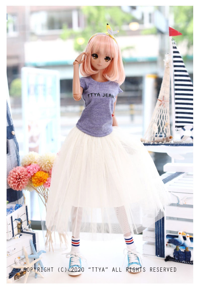 K&ouml;nnte beinhalten: Eine Puppe tr&auml;gt ein lila T-Shirt mit dem Aufdruck "TTYA JEAN", einen wei&szlig;en T&uuml;llrock, blau-rot gestreifte Socken und blaue Turnschuhe. Die Puppe hat rosa Haare und blaue Augen.