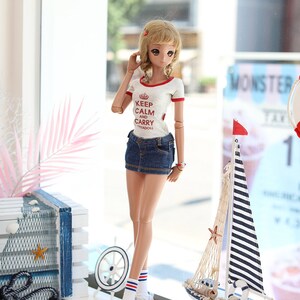 Puede incluir: Una mu&ntilde;eca con una camiseta blanca con texto rojo que dice "Keep Calm and Carry" y una minifalda vaquera azul. La mu&ntilde;eca lleva zapatillas blancas con rayas rojas y azules en los calcetines. La mu&ntilde;eca est&aacute; de pie frente a una ventana con una decoraci&oacute;n de velero en el alf&eacute;izar de la ventana.