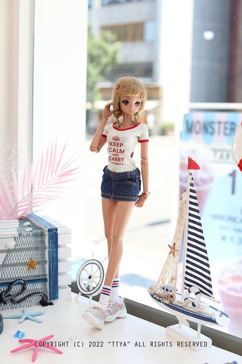 Puede incluir: Una mu&ntilde;eca con una camiseta blanca con texto rojo que dice "Keep Calm and Carry" y una minifalda vaquera azul. La mu&ntilde;eca lleva zapatillas blancas con rayas rojas y azules en los calcetines. La mu&ntilde;eca est&aacute; de pie frente a una ventana con una decoraci&oacute;n de velero en el alf&eacute;izar de la ventana.
