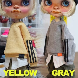 May include: Two dolls wearing miniature sweatshirts. One doll is in a yellow sweatshirt with black and gold stripes on the sleeves. The other doll is in a gray sweatshirt with black stripes on the sleeves. The word "YELLOW" is below the yellow sweatshirt and "GRAY" is below the gray sweatshirt.