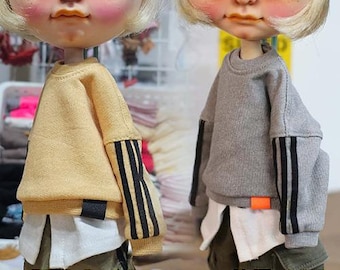 Puede incluir: Dos muñecas con sudaderas en miniatura. Una muñeca lleva una sudadera amarilla con rayas negras y doradas en las mangas. La otra muñeca lleva una sudadera gris con rayas negras en las mangas. Las palabras "YELLOW" y "GRAY" están debajo.