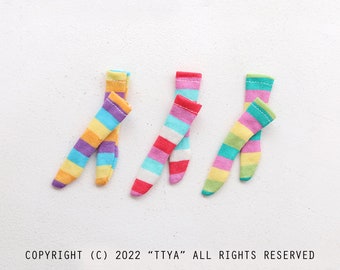 Puede incluir: Tres pares de calcetines pequeños a rayas en colores vibrantes. El primer par presenta rayas amarillas, naranjas y moradas. El segundo par tiene rayas rojas, blancas y turquesas. El tercer par muestra rayas verdes, amarillas y rosas. COPYRIGHT (C) 2022 “TTYA” ALL RIGHTS RESERVED