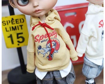 Puede incluir: Una muñeca con una sudadera con capucha amarilla con un gráfico de lobo y el texto "Speedy Wolf". La muñeca también lleva pantalones cortos de mezclilla, zapatillas deportivas y una gorra de béisbol roja. Un letrero amarillo de límite de velocidad está en el fondo.