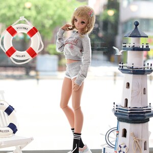 May include: A doll wearing a grey sweatshirt with the words "New York" on it, grey shorts, black and white striped socks, and pink and blue sneakers. The doll is standing in front of a window with a life preserver that says "Welcome Aboard" hanging on it. There is a lighthouse model to the right of the doll.