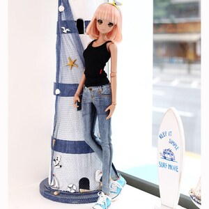 Puede incluir: Una mu&ntilde;eca con el pelo rosa que lleva un top negro sin mangas, pantalones vaqueros azules y zapatillas azules. La mu&ntilde;eca est&aacute; de pie junto a un faro en miniatura y una tabla de surf con el texto "Keep it simple, surf more".