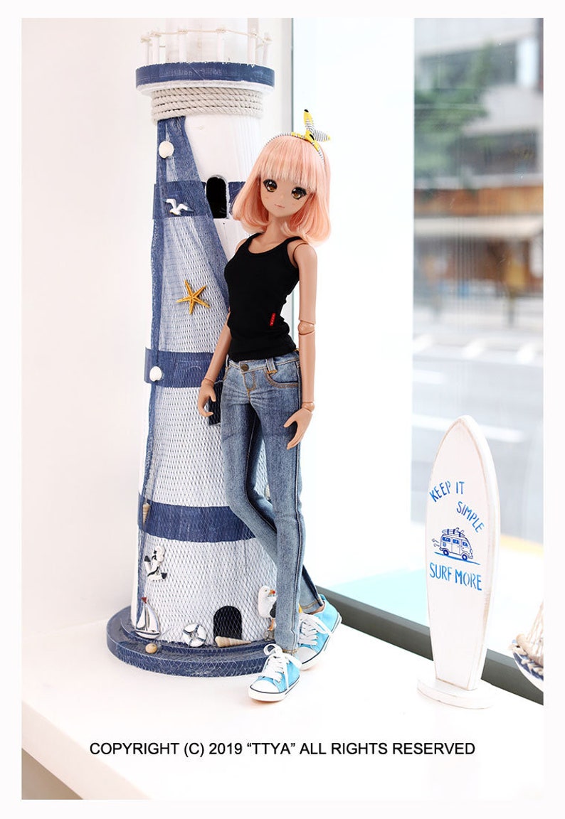 Puede incluir: Una mu&ntilde;eca con el pelo rosa que lleva un top negro sin mangas, pantalones vaqueros azules y zapatillas azules. La mu&ntilde;eca est&aacute; de pie junto a un faro en miniatura y una tabla de surf con el texto "Keep it simple, surf more".