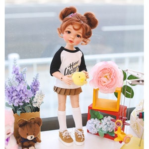 Puede incluir: Una mu&ntilde;eca con el pelo casta&ntilde;o en coletas que lleva una camiseta de b&eacute;isbol blanca y negra con el texto "Honor Victor" en ella, una falda marr&oacute;n, calcetines blancos y zapatos marrones. La mu&ntilde;eca sostiene un juguete amarillo de pollito. La mu&ntilde;eca est&aacute; de pie sobre una superficie blanca con un oso de peluche marr&oacute;n, un ramo de flores moradas y una flor rosa.