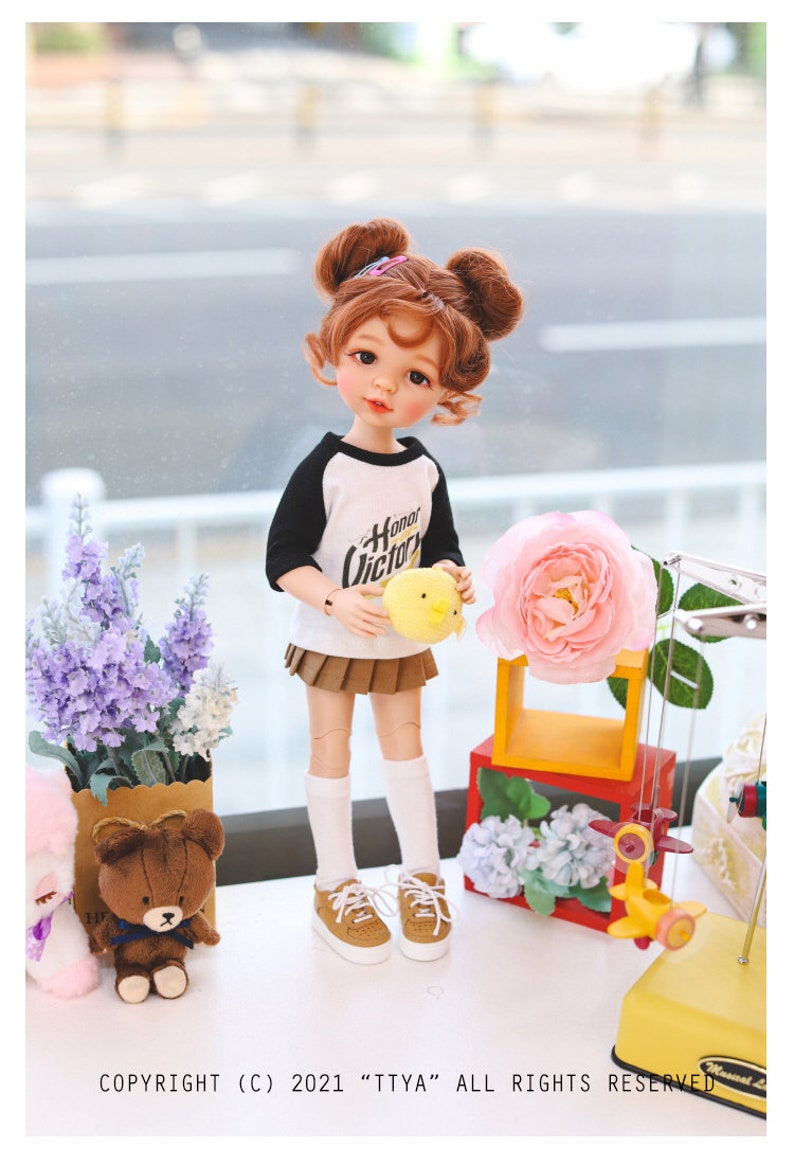 Puede incluir: Una mu&ntilde;eca con el pelo casta&ntilde;o en coletas que lleva una camiseta de b&eacute;isbol blanca y negra con el texto "Honor Victor" en ella, una falda marr&oacute;n, calcetines blancos y zapatos marrones. La mu&ntilde;eca sostiene un juguete amarillo de pollito. La mu&ntilde;eca est&aacute; de pie sobre una superficie blanca con un oso de peluche marr&oacute;n, un ramo de flores moradas y una flor rosa.