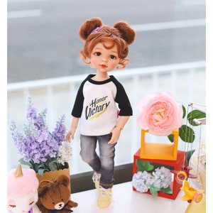 Puede incluir: Una mu&ntilde;eca con el pelo casta&ntilde;o en coletas que lleva una camiseta de b&eacute;isbol blanca y negra con las palabras "Honor Victory" impresas. La mu&ntilde;eca lleva pantalones vaqueros azules y zapatillas blancas. La mu&ntilde;eca est&aacute; de pie sobre una superficie blanca con un juguete de peluche de unicornio, un juguete de peluche de oso marr&oacute;n, un ramo de lavanda, una flor rosa y un avi&oacute;n de juguete amarillo.