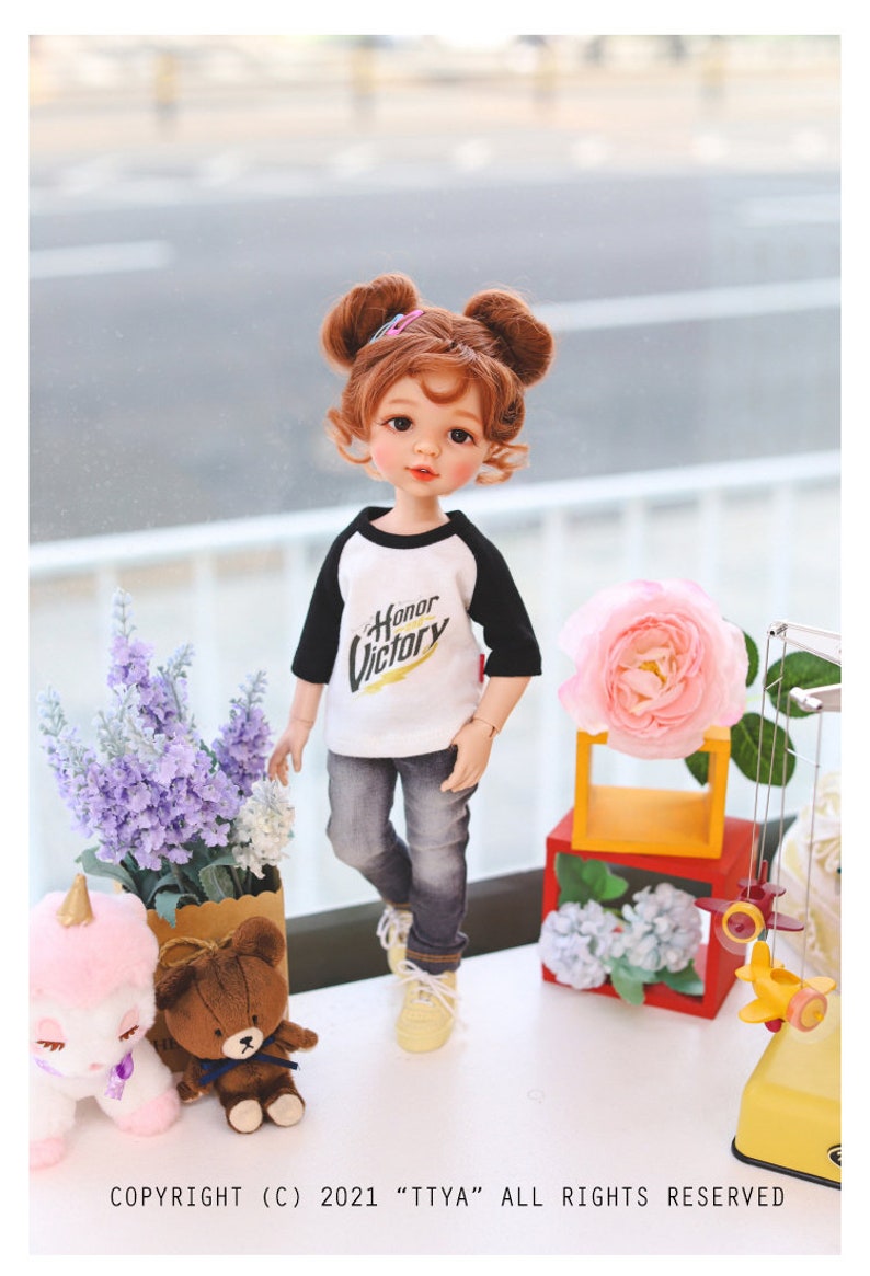 Puede incluir: Una mu&ntilde;eca con el pelo casta&ntilde;o en coletas que lleva una camiseta de b&eacute;isbol blanca y negra con las palabras "Honor Victory" impresas. La mu&ntilde;eca lleva pantalones vaqueros azules y zapatillas blancas. La mu&ntilde;eca est&aacute; de pie sobre una superficie blanca con un juguete de peluche de unicornio, un juguete de peluche de oso marr&oacute;n, un ramo de lavanda, una flor rosa y un avi&oacute;n de juguete amarillo.