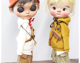 Puede incluir: Dos muñecas personalizadas vestidas con ropa urbana. Una muñeca lleva un gorro naranja, una sudadera con capucha blanca y pantalones cortos color caqui. La otra muñeca lleva una sudadera con capucha amarilla y pantalones cortos color caqui. Ambas muñecas tienen zapatillas y bolsos pequeños. El texto "TTYA.COM" es visible.