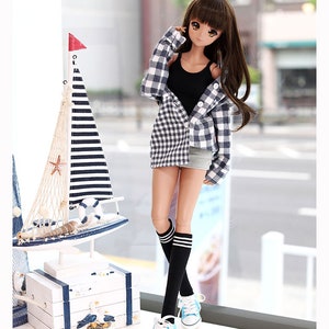 Puede incluir: Una mu&ntilde;eca con un top negro sin mangas, una camisa de cuadros azul y blanco, una falda vaquera, calcetines negros hasta la rodilla con rayas blancas y zapatillas azules. La mu&ntilde;eca tiene el pelo largo y casta&ntilde;o y los ojos marrones. La mu&ntilde;eca est&aacute; de pie frente a una ventana con una decoraci&oacute;n de velero de madera blanca en un estante debajo.