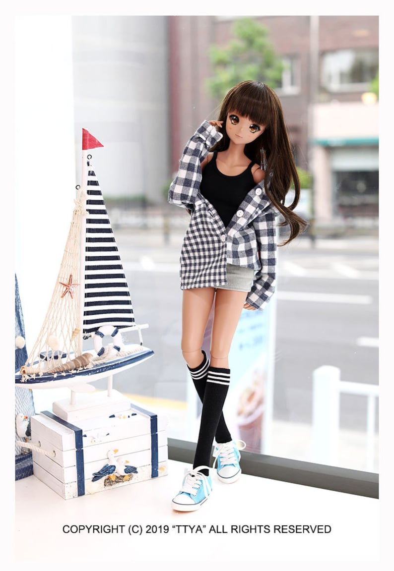 Puede incluir: Una mu&ntilde;eca con un top negro sin mangas, una camisa de cuadros azul y blanco, una falda vaquera, calcetines negros hasta la rodilla con rayas blancas y zapatillas azules. La mu&ntilde;eca tiene el pelo largo y casta&ntilde;o y los ojos marrones. La mu&ntilde;eca est&aacute; de pie frente a una ventana con una decoraci&oacute;n de velero de madera blanca en un estante debajo.