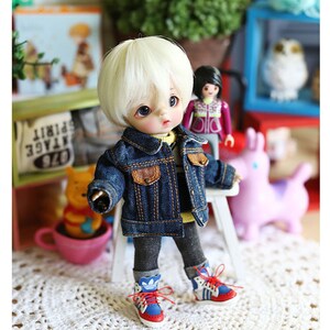 以下が含まれることがあります： 金髪のウィッグをつけた人形が、青いデニムジャケット、グレーのパンツ、赤と青のスニーカーを履いています。人形は白いレースのドイリーの上に座っています。