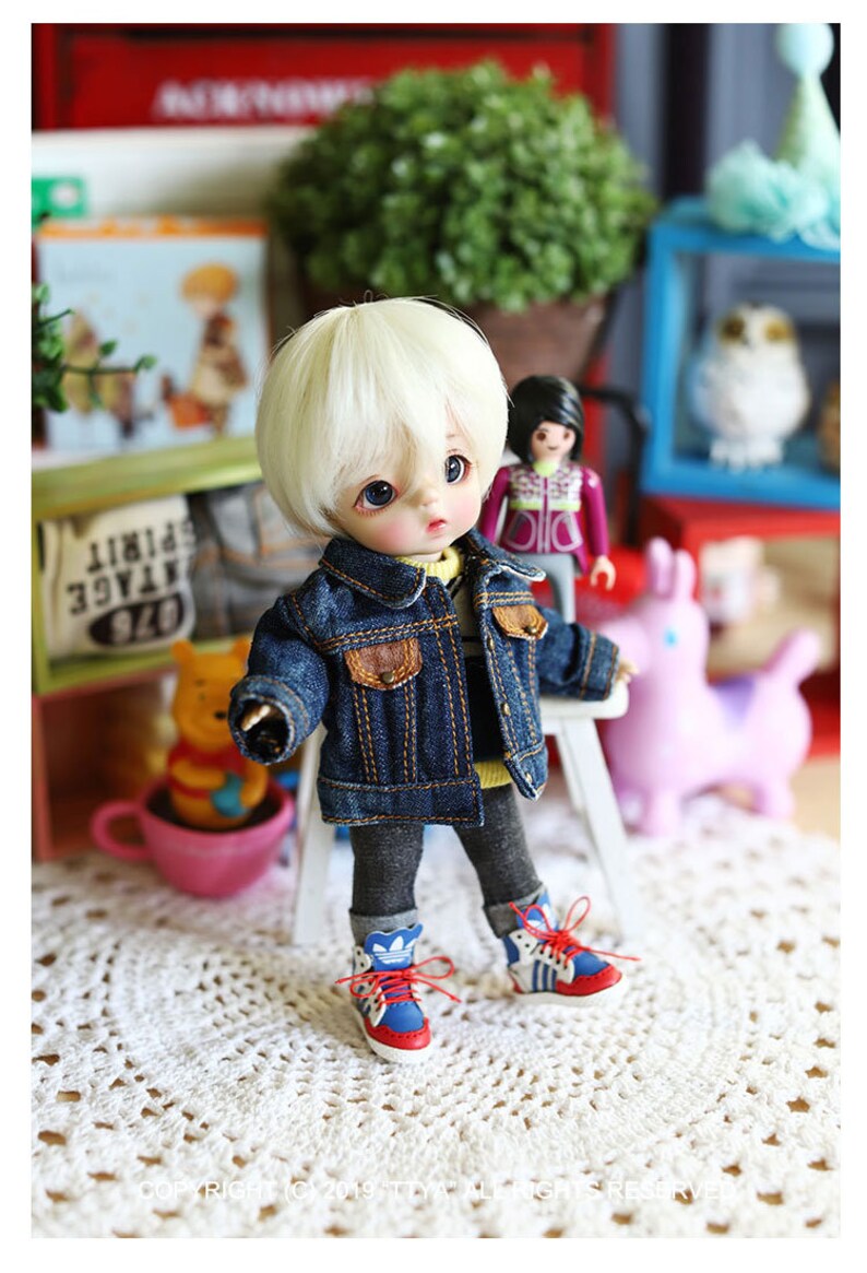 以下が含まれることがあります： 金髪のウィッグをつけた人形が、青いデニムジャケット、グレーのパンツ、赤と青のスニーカーを履いています。人形は白いレースのドイリーの上に座っています。