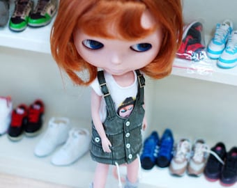 May include: A red-haired doll wearing a green denim overall dress with a white t-shirt underneath. The doll is wearing gray sneakers with green accents and has blue eyes. The doll is standing on a wooden floor in front of a shelf with various shoes.