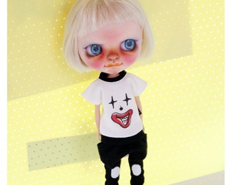 Puede incluir: Una muñeca con cabello rubio que lleva una camiseta blanca con un diseño de cara de payaso rojo y negro, pantalones negros con detalles blancos y zapatos negros. La muñeca está de pie sobre una superficie blanca con un fondo amarillo.