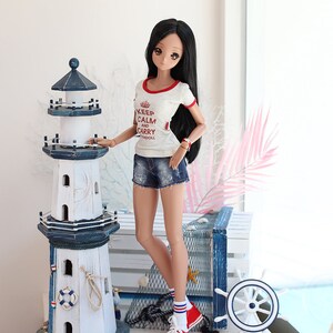 Puede incluir: Una mu&ntilde;eca con una camiseta blanca con ribetes rojos y el texto "Keep Calm and Carry Madoll" en rojo. La mu&ntilde;eca lleva pantalones cortos de mezclilla azul y calcetines a rayas rojas y blancas con zapatillas rojas. La mu&ntilde;eca est&aacute; de pie junto a un modelo de faro blanco y azul con un salvavidas y una cuerda.