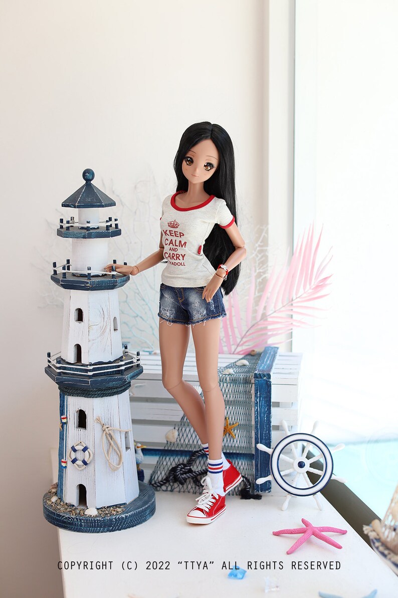 Puede incluir: Una mu&ntilde;eca con una camiseta blanca con ribetes rojos y el texto "Keep Calm and Carry Madoll" en rojo. La mu&ntilde;eca lleva pantalones cortos de mezclilla azul y calcetines a rayas rojas y blancas con zapatillas rojas. La mu&ntilde;eca est&aacute; de pie junto a un modelo de faro blanco y azul con un salvavidas y una cuerda.