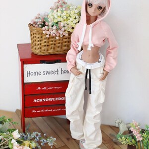 Puede incluir: Una mu&ntilde;eca con una sudadera rosa con capucha recortada, un cord&oacute;n blanco y pantalones blancos. La mu&ntilde;eca tiene el pelo largo y blanco y sombra de ojos rosa. La mu&ntilde;eca est&aacute; de pie frente a una c&oacute;moda de madera roja con cajones que tienen texto. El texto en los cajones dice "Home sweet home", "In all your ways" y "Acknowledge".