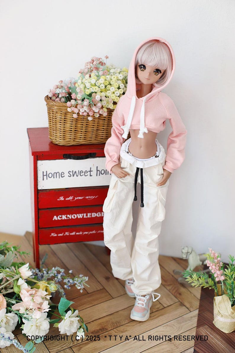 Puede incluir: Una mu&ntilde;eca con una sudadera rosa con capucha recortada, un cord&oacute;n blanco y pantalones blancos. La mu&ntilde;eca tiene el pelo largo y blanco y sombra de ojos rosa. La mu&ntilde;eca est&aacute; de pie frente a una c&oacute;moda de madera roja con cajones que tienen texto. El texto en los cajones dice "Home sweet home", "In all your ways" y "Acknowledge".