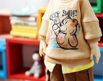 May include: A beige long-sleeve sweater with a ribbed crew neck and cuffs, featuring a graphic of a bear and the text "KEEP SMILE" in black, orange, and blue. The sweater is layered over a white and yellow striped shirt and brown cargo shorts.
