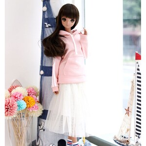 K&ouml;nnte beinhalten: Eine Puppe tr&auml;gt ein rosa Hoodie mit wei&szlig;en Kordeln, einen wei&szlig;en T&uuml;llrock und blau-wei&szlig; gestreifte Socken. Die Puppe steht neben einem Miniatur-Leuchtturm und einem Segelboot.