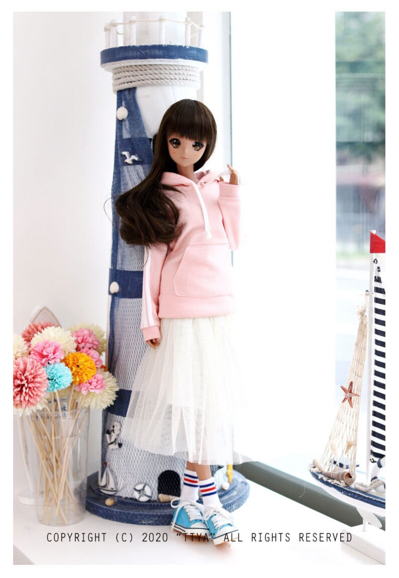 K&ouml;nnte beinhalten: Eine Puppe tr&auml;gt ein rosa Hoodie mit wei&szlig;en Kordeln, einen wei&szlig;en T&uuml;llrock und blau-wei&szlig; gestreifte Socken. Die Puppe steht neben einem Miniatur-Leuchtturm und einem Segelboot.