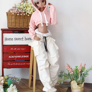 Puede incluir: Una sudadera con capucha rosa recortada con un cord&oacute;n blanco y un ribete blanco alrededor del dobladillo. La sudadera la lleva una mu&ntilde;eca con pantalones blancos y cintur&oacute;n negro. La mu&ntilde;eca est&aacute; de pie sobre un suelo de madera con un taburete de madera y una cesta de mimbre llena de flores.