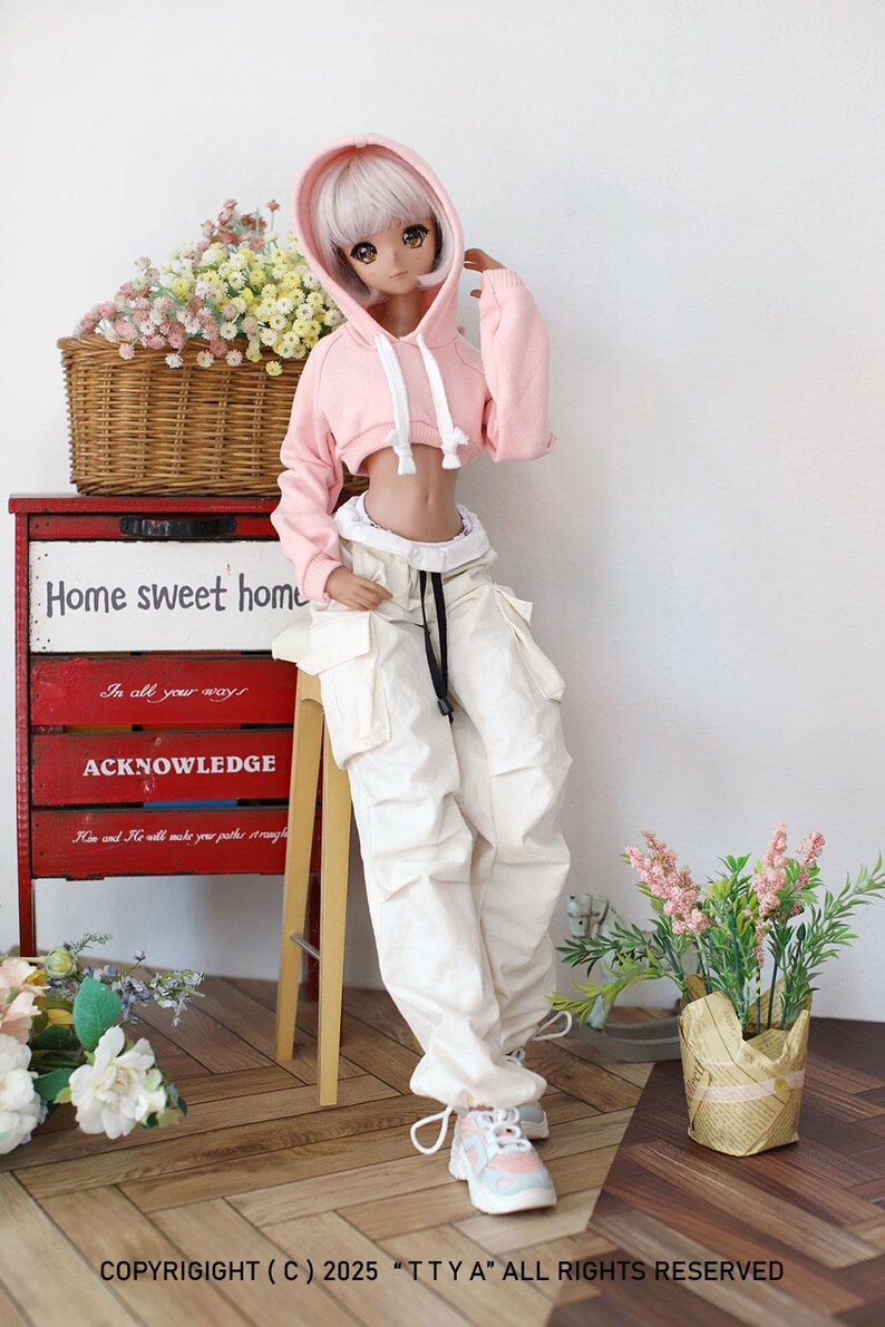 Puede incluir: Una sudadera con capucha rosa recortada con un cord&oacute;n blanco y un ribete blanco alrededor del dobladillo. La sudadera la lleva una mu&ntilde;eca con pantalones blancos y cintur&oacute;n negro. La mu&ntilde;eca est&aacute; de pie sobre un suelo de madera con un taburete de madera y una cesta de mimbre llena de flores.