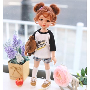 Puede incluir: Una mu&ntilde;eca con el pelo rojo en coletas que lleva una camiseta de b&eacute;isbol blanca y negra con las palabras "Honor Victory" impresas. La mu&ntilde;eca lleva un osito de peluche marr&oacute;n y lleva pantalones cortos de mezclilla azul, calcetines blancos y zapatos marrones. La mu&ntilde;eca est&aacute; de pie sobre una superficie blanca con una flor rosa, un caballo de balanc&iacute;n de madera y dos pollitos amarillos.