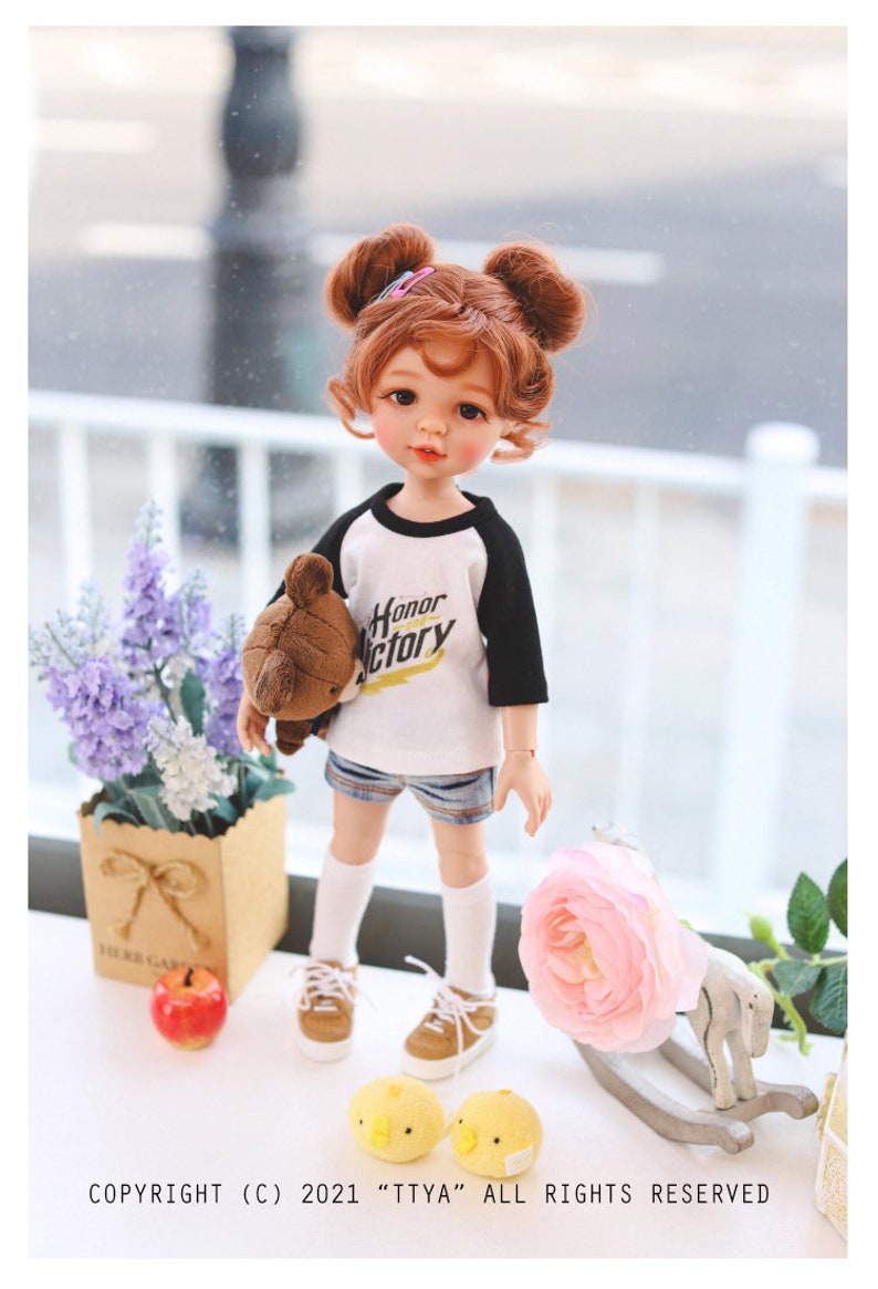 Puede incluir: Una mu&ntilde;eca con el pelo rojo en coletas que lleva una camiseta de b&eacute;isbol blanca y negra con las palabras "Honor Victory" impresas. La mu&ntilde;eca lleva un osito de peluche marr&oacute;n y lleva pantalones cortos de mezclilla azul, calcetines blancos y zapatos marrones. La mu&ntilde;eca est&aacute; de pie sobre una superficie blanca con una flor rosa, un caballo de balanc&iacute;n de madera y dos pollitos amarillos.