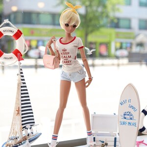 Puede incluir: Una mu&ntilde;eca que lleva una camiseta blanca con ribetes rojos y el texto "Keep Calm and Carry Yadoll" en rojo. La mu&ntilde;eca tambi&eacute;n lleva pantalones cortos de mezclilla, calcetines blancos con rayas rojas y azules y zapatos rosas. La mu&ntilde;eca sostiene un bolso de mano rosa. La mu&ntilde;eca est&aacute; de pie frente a una ventana con un salvavidas y un velero en el fondo. La mu&ntilde;eca est&aacute; de pie sobre una superficie blanca con una tabla de surf y una caja de madera en el fondo. La tabla de surf tiene el texto "Keep It Simple Surf More" en azul.