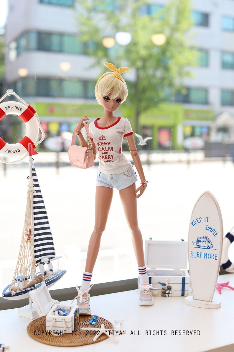 Puede incluir: Una mu&ntilde;eca que lleva una camiseta blanca con ribetes rojos y el texto "Keep Calm and Carry Yadoll" en rojo. La mu&ntilde;eca tambi&eacute;n lleva pantalones cortos de mezclilla, calcetines blancos con rayas rojas y azules y zapatos rosas. La mu&ntilde;eca sostiene un bolso de mano rosa. La mu&ntilde;eca est&aacute; de pie frente a una ventana con un salvavidas y un velero en el fondo. La mu&ntilde;eca est&aacute; de pie sobre una superficie blanca con una tabla de surf y una caja de madera en el fondo. La tabla de surf tiene el texto "Keep It Simple Surf More" en azul.