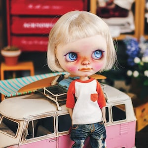 May include: A doll with blonde hair and blue eyes wearing a red and white striped shirt, blue jeans, and blue sneakers. The doll is standing next to a pink and white toy van with a surfboard on top.