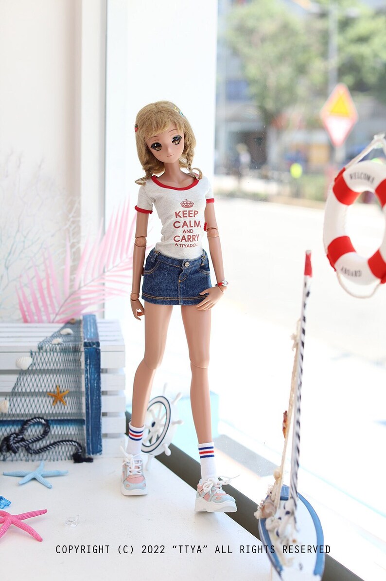 Puede incluir: Una mu&ntilde;eca que lleva una camiseta blanca con ribetes rojos y el texto "Keep Calm and Carry On" en rojo. La mu&ntilde;eca tambi&eacute;n lleva una falda vaquera, calcetines blancos con rayas rojas y azules y zapatillas blancas. La mu&ntilde;eca est&aacute; de pie frente a una ventana con un salvavidas y un velero modelo visible en el exterior.
