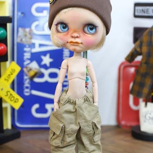 May include: A doll with a brown beanie featuring a "Smile" patch, blue eyes, and rosy cheeks. The doll wears tan cargo pants and black and white sneakers. The doll's arms and torso are visible. The background includes a blue license plate and a traffic signal.