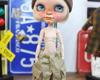 Puede incluir: Una muñeca con un gorro marrón con un parche de "Smile", ojos azules y mejillas rosadas. La muñeca lleva pantalones cargo color beige y zapatillas deportivas blancas y negras. Se ven los brazos y el torso de la muñeca. El fondo incluye una placa azul y un semáforo.
