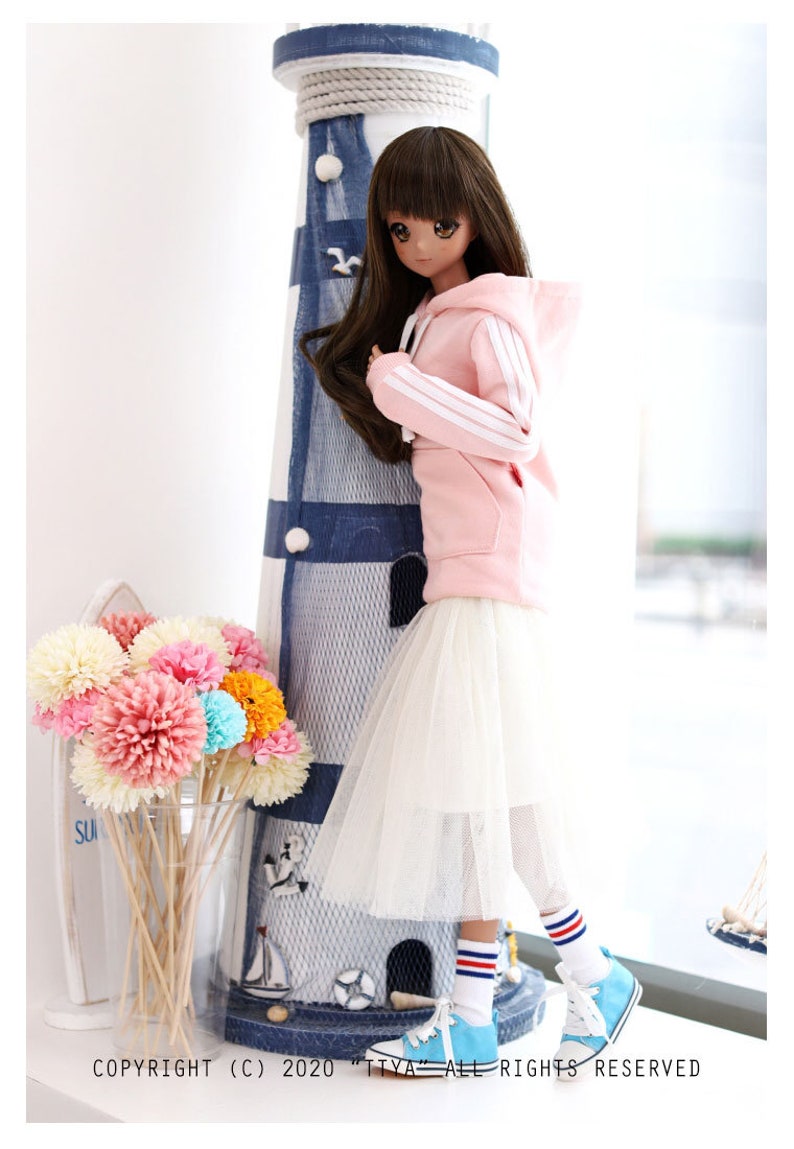 K&ouml;nnte beinhalten: Ein rosa Hoodie mit wei&szlig;en Streifen an den &Auml;rmeln, ein wei&szlig;er T&uuml;llrock und blau-wei&szlig; gestreifte Socken. Die Puppe tr&auml;gt blaue Turnschuhe und steht neben einem blau-wei&szlig;en Leuchtturm.